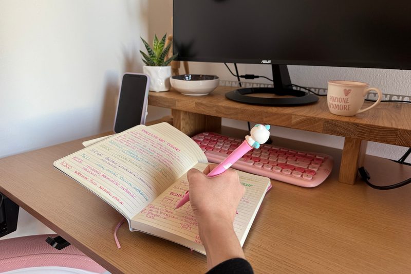 Una mano scrive appunti con la penna rosa su un quaderno, accanto alla tastiera rosa